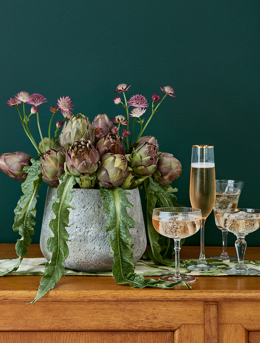 Coupes et flûte de vin pétillant rosé près d'un bouquet de fleurs d'astrances et artichauts, fond vert forêt
